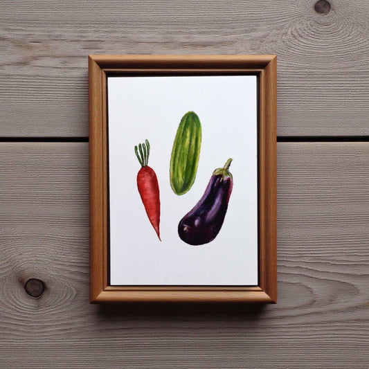 Framed artwork of a red carrot, green cucumber, and purple eggplant on a wooden surface.
