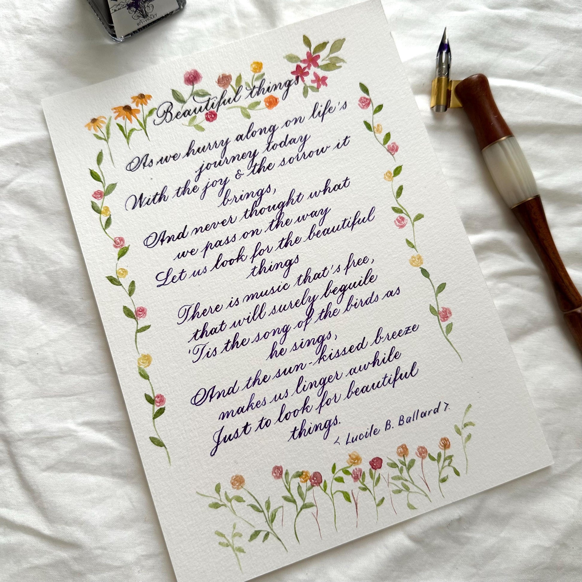Handwritten calligraphy letter with watercolor floral border and dip pen beside it