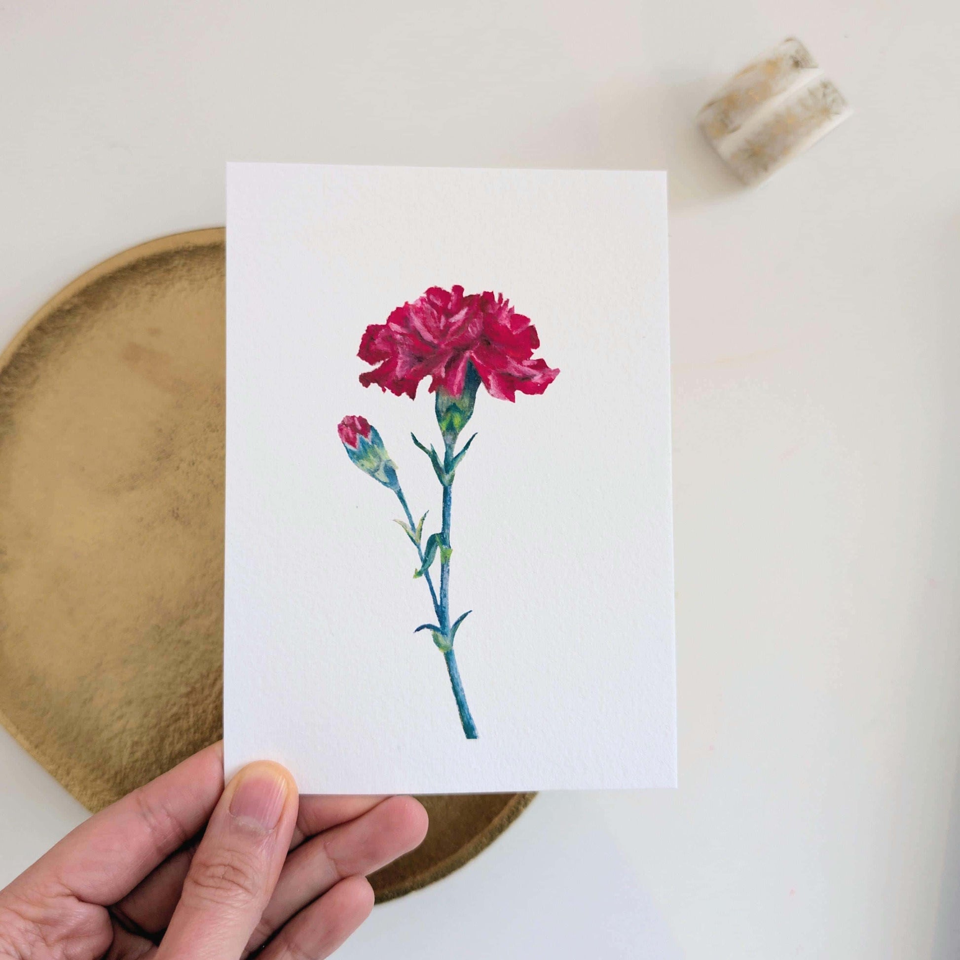 Angled handheld view of January birth month carnation postcard, oil pastel floral artwork on A6 fine art card, HerPens