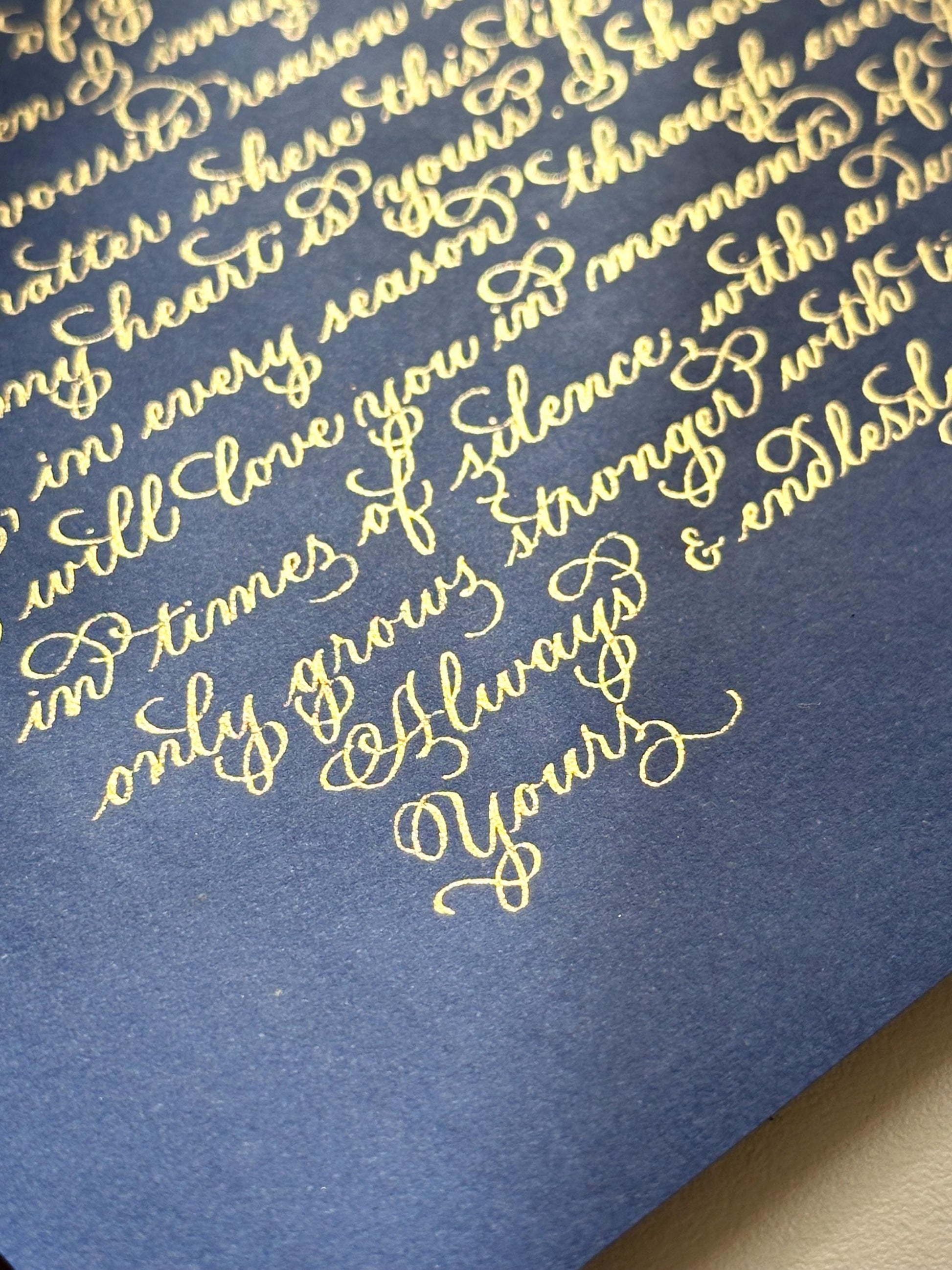 Handwritten calligraphy letter on navy paper in gold ink captured at angle with rich contrast elegant script herpens