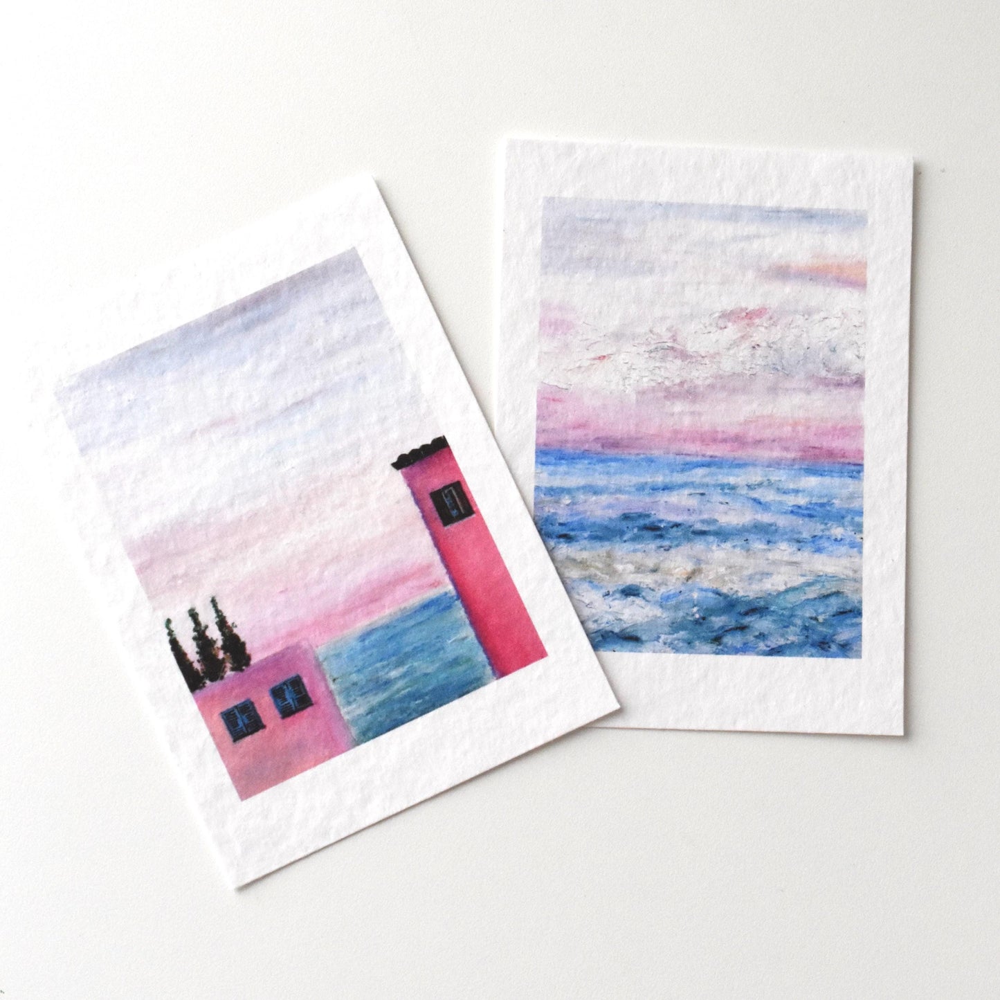 Close-up of two oil pastel landscape postcards by HerPens — pink seaside house and pastel ocean sunset, printed on textured fine art paper.