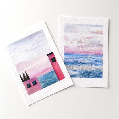Close-up of two oil pastel landscape postcards by HerPens — pink seaside house and pastel ocean sunset, printed on textured fine art paper.