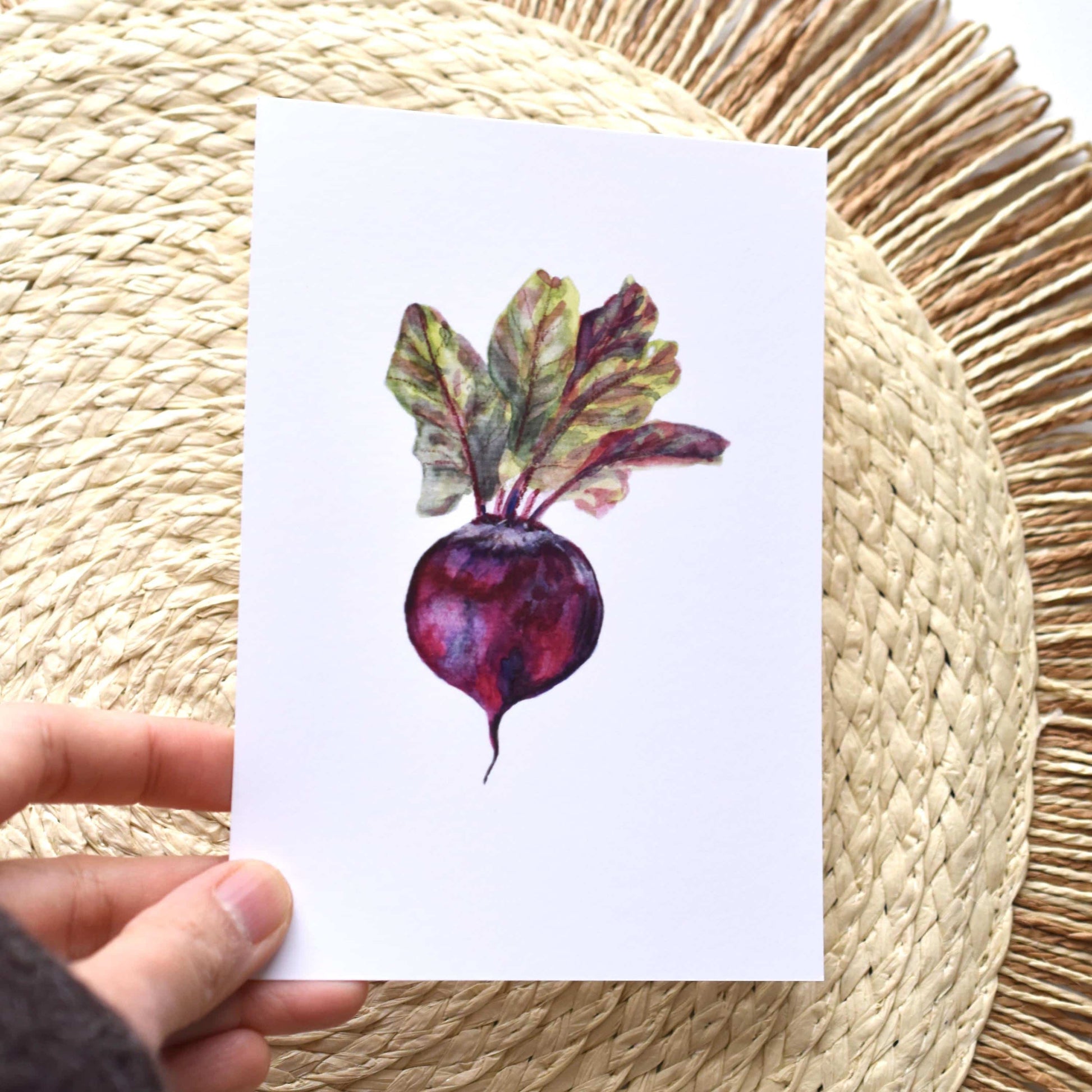 Hand holding a card with a watercolor illustration of a beetroot on a woven mat background