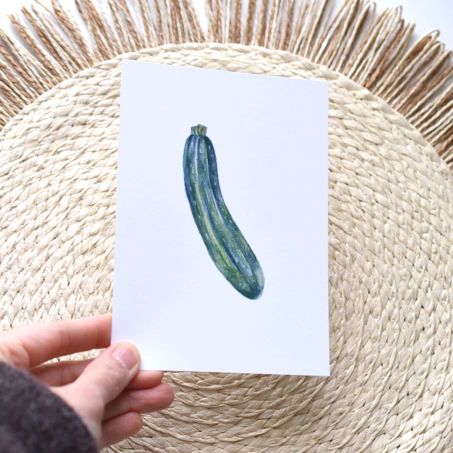 Hand holding a card with a zucchini illustration on a woven mat