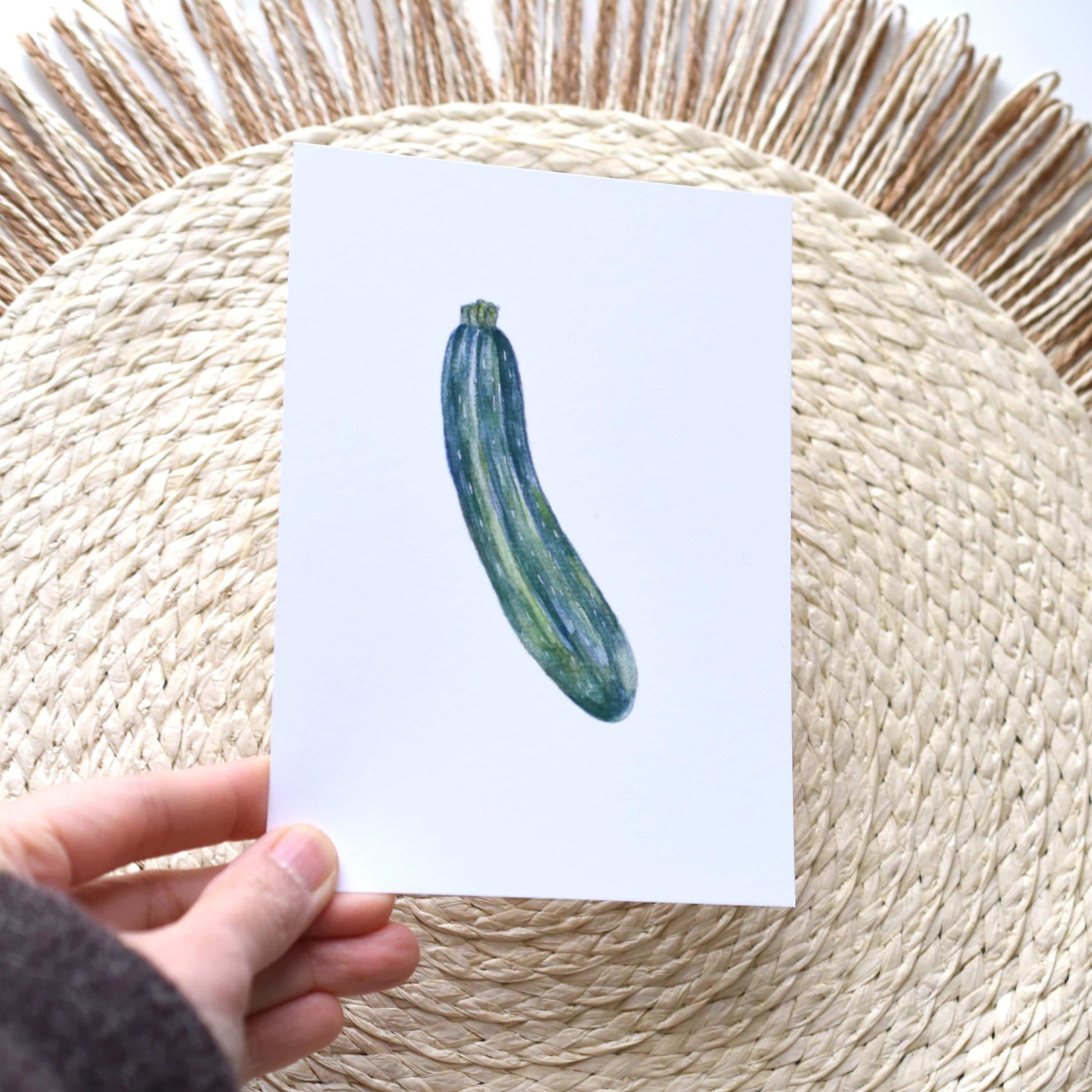 Hand holding a card with a zucchini illustration on a woven mat