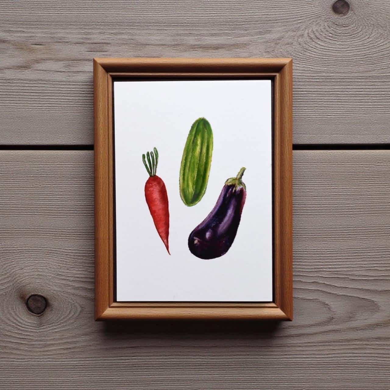 Framed artwork of a red carrot, green cucumber, and purple eggplant on a wooden surface.