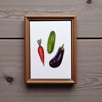 Framed artwork of a red carrot, green cucumber, and purple eggplant on a wooden surface.