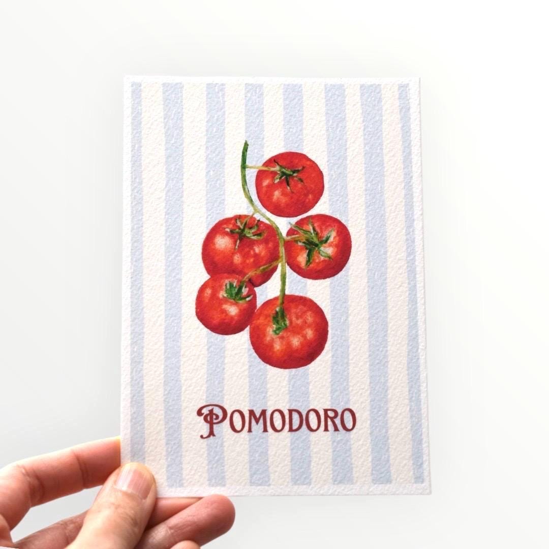 Hand holding the “Pomodoro” A6 postcard — red vine tomatoes painted in oil pastel over light blue stripes with the word Pomodoro, printed on thick fine art paper by HerPens.