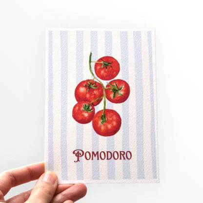 Hand holding the “Pomodoro” A6 postcard — red vine tomatoes painted in oil pastel over light blue stripes with the word Pomodoro, printed on thick fine art paper by HerPens.