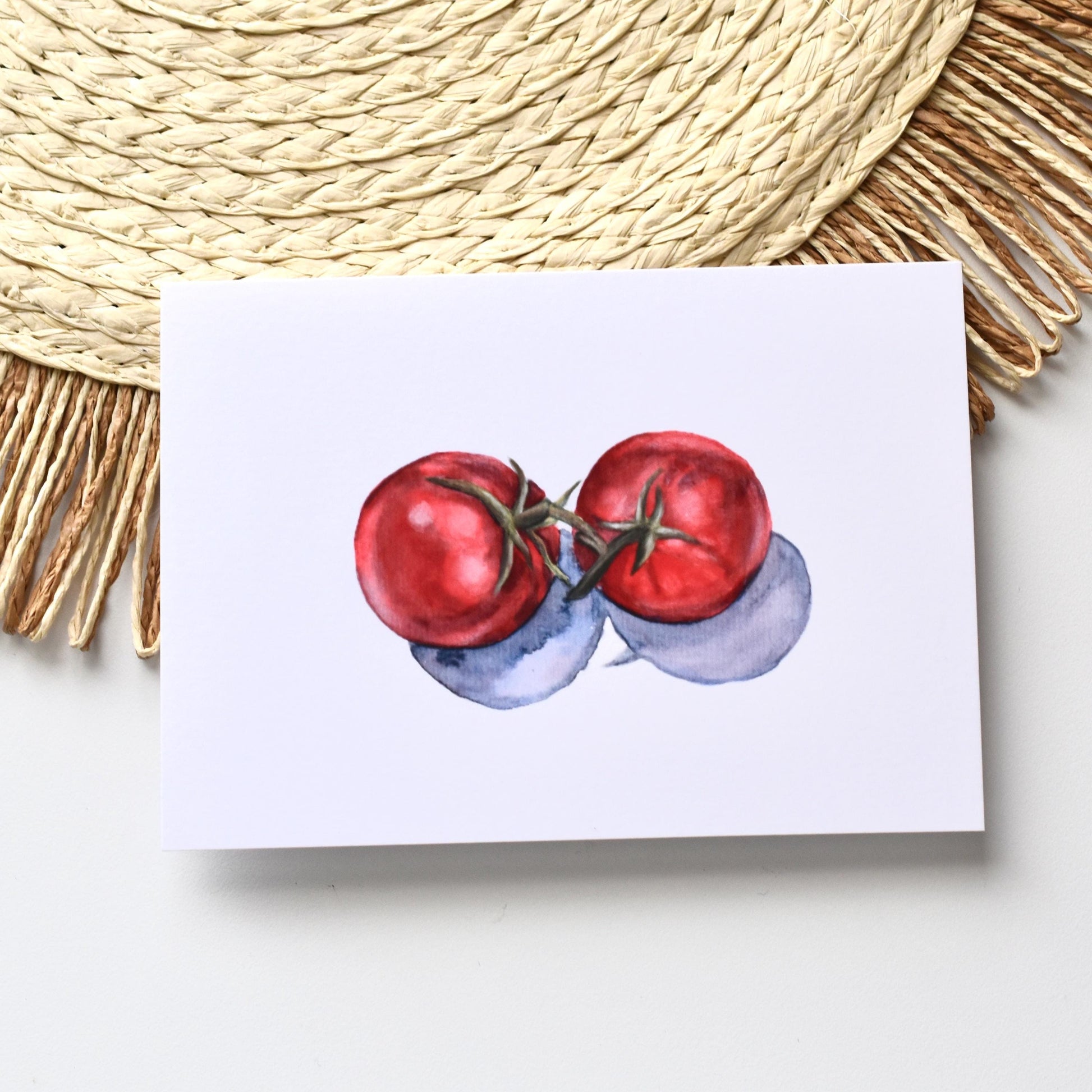 A6 watercolour tomato postcard placed on a woven mat, featuring two red tomatoes with soft painterly shading, HerPens