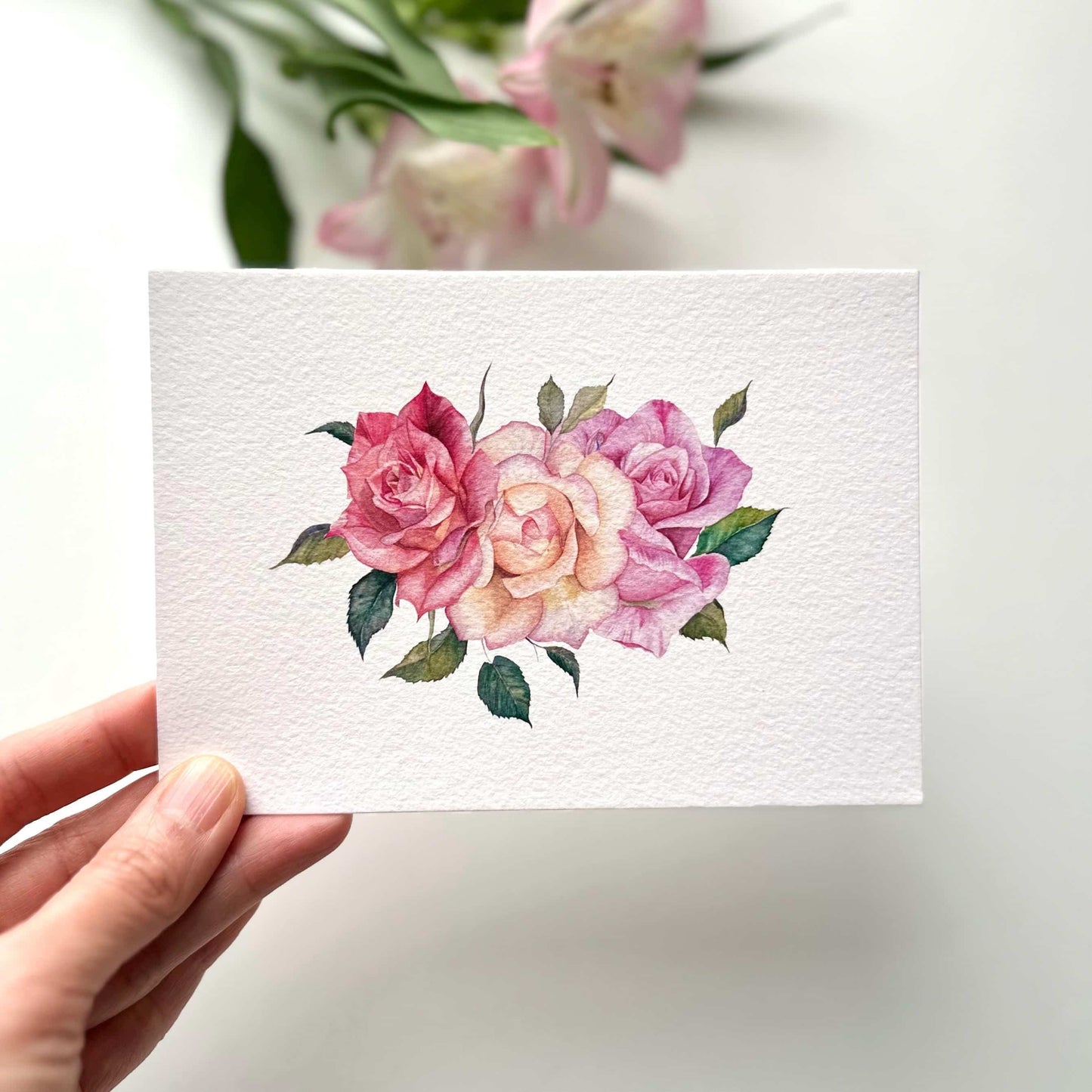Hand holding A6 watercolor roses postcard printed on textured fine art paper, soft floral illustration with gentle rose details, HerPens